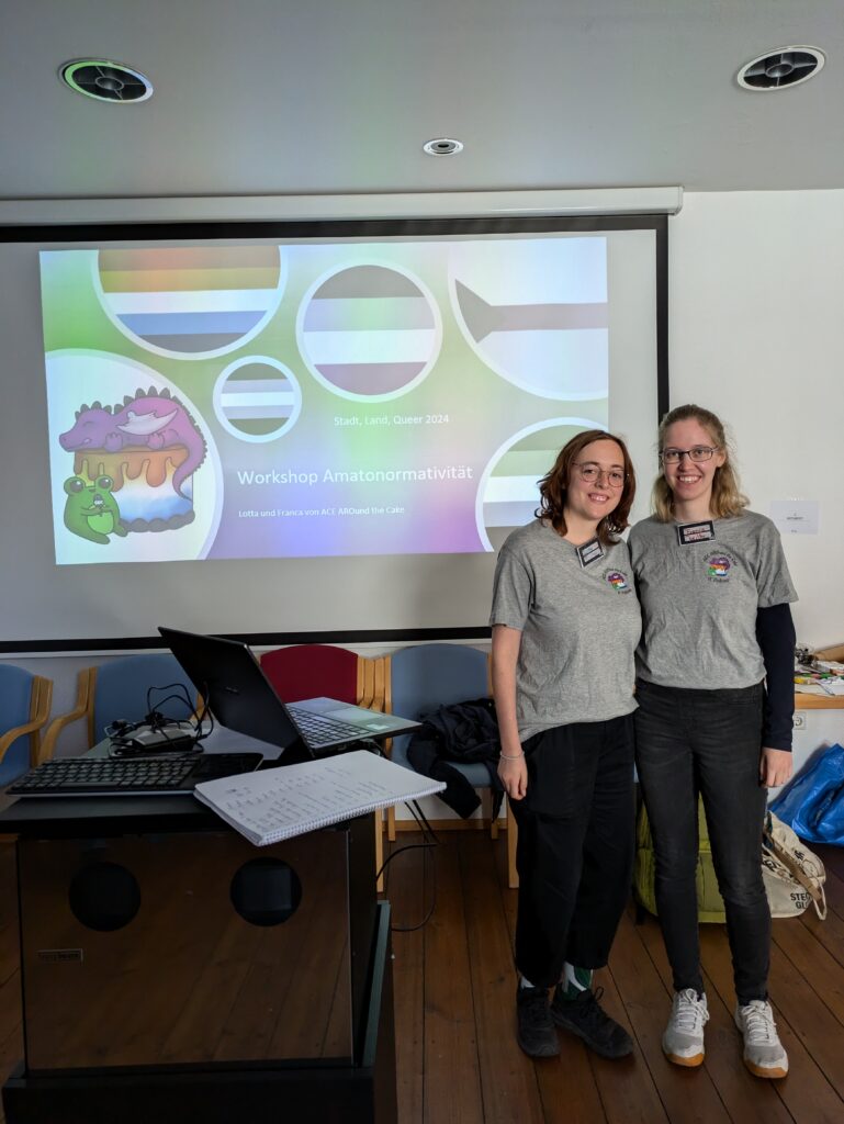 Franca und Lotta aus dem Podcast-Team stehen vor einer Leinwand mit der Titelfolie ihres Workshops zum Thema Amatonormativität bei Stadt, Land, Queer 2024