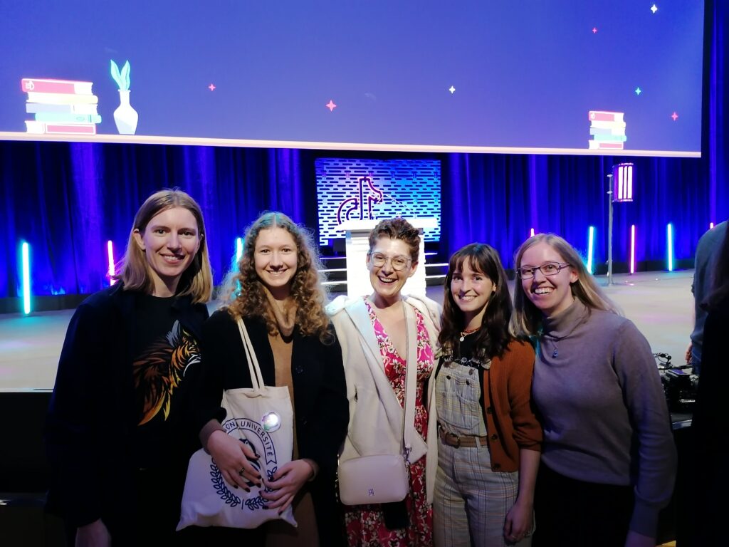 Fünf Personen stehen vor einer blau beleuchteten Bühne mit großer Leinwand und Rednerpult. Von links nach rechts: Lea, Clara, Jana Crämer, Hannah und Franca